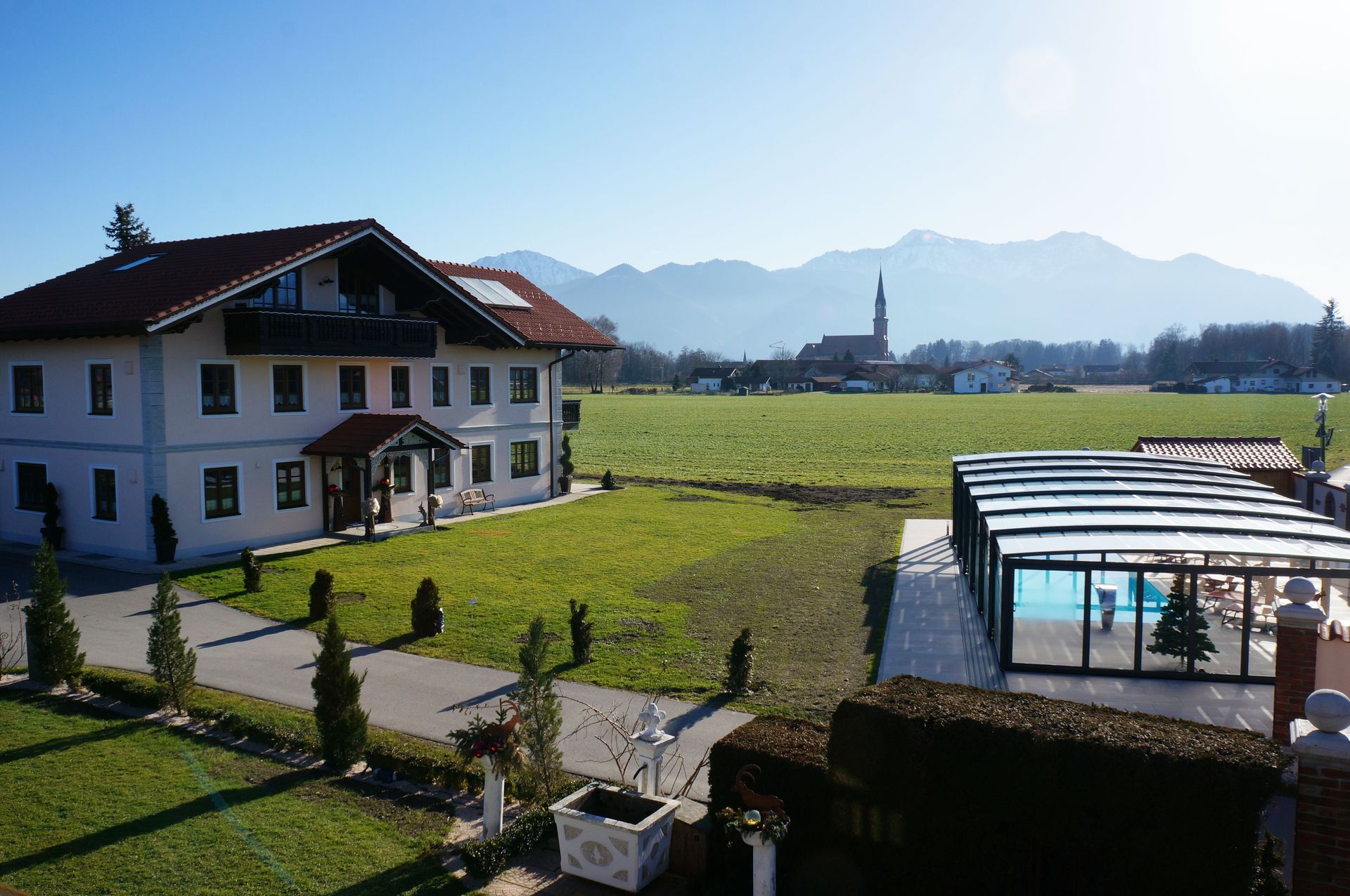 Großes weißes Landhaus mit rotem Dach neben überdachtem Schwimmbecken, im Hintergrund Berge und Kirchturm